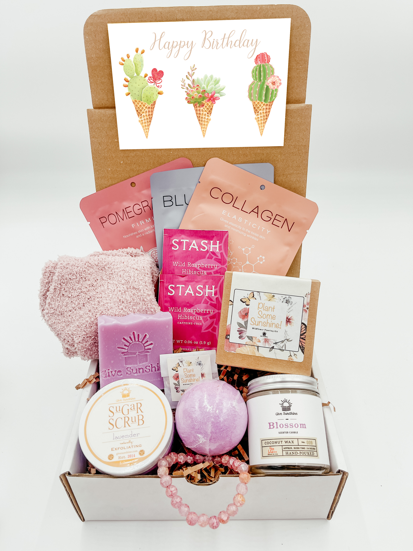 A gift box with various pink and purple colored items including a glass beaded bracelet, comfy socks, a blossoms-scented candle, lavender sugar scrub, a bath bomb, lavender goats milk soap, a wildflower kit, and a packet of wild raspberry hibiscus tea.