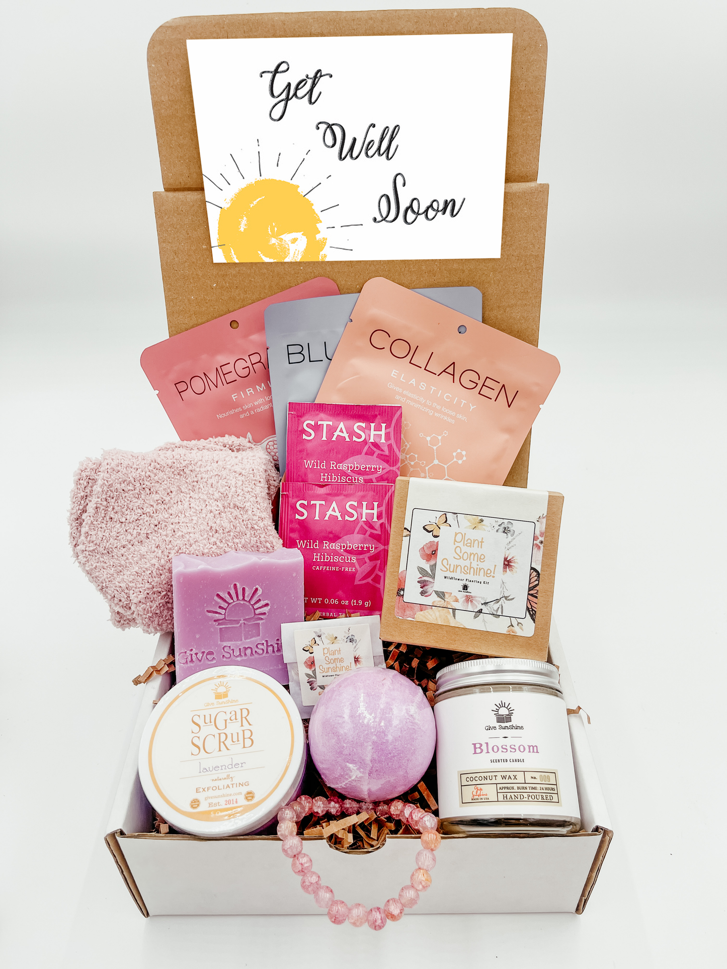 A gift box with various pink and purple colored items including a glass beaded bracelet, comfy socks, a blossoms-scented candle, lavender sugar scrub, a bath bomb, lavender goats milk soap, a wildflower kit, and a packet of wild raspberry hibiscus tea.