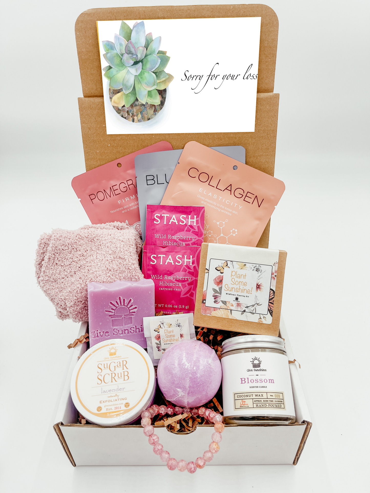 A gift box with various pink and purple colored items including a glass beaded bracelet, comfy socks, a blossoms-scented candle, lavender sugar scrub, a bath bomb, lavender goats milk soap, a wildflower kit, and a packet of wild raspberry hibiscus tea.