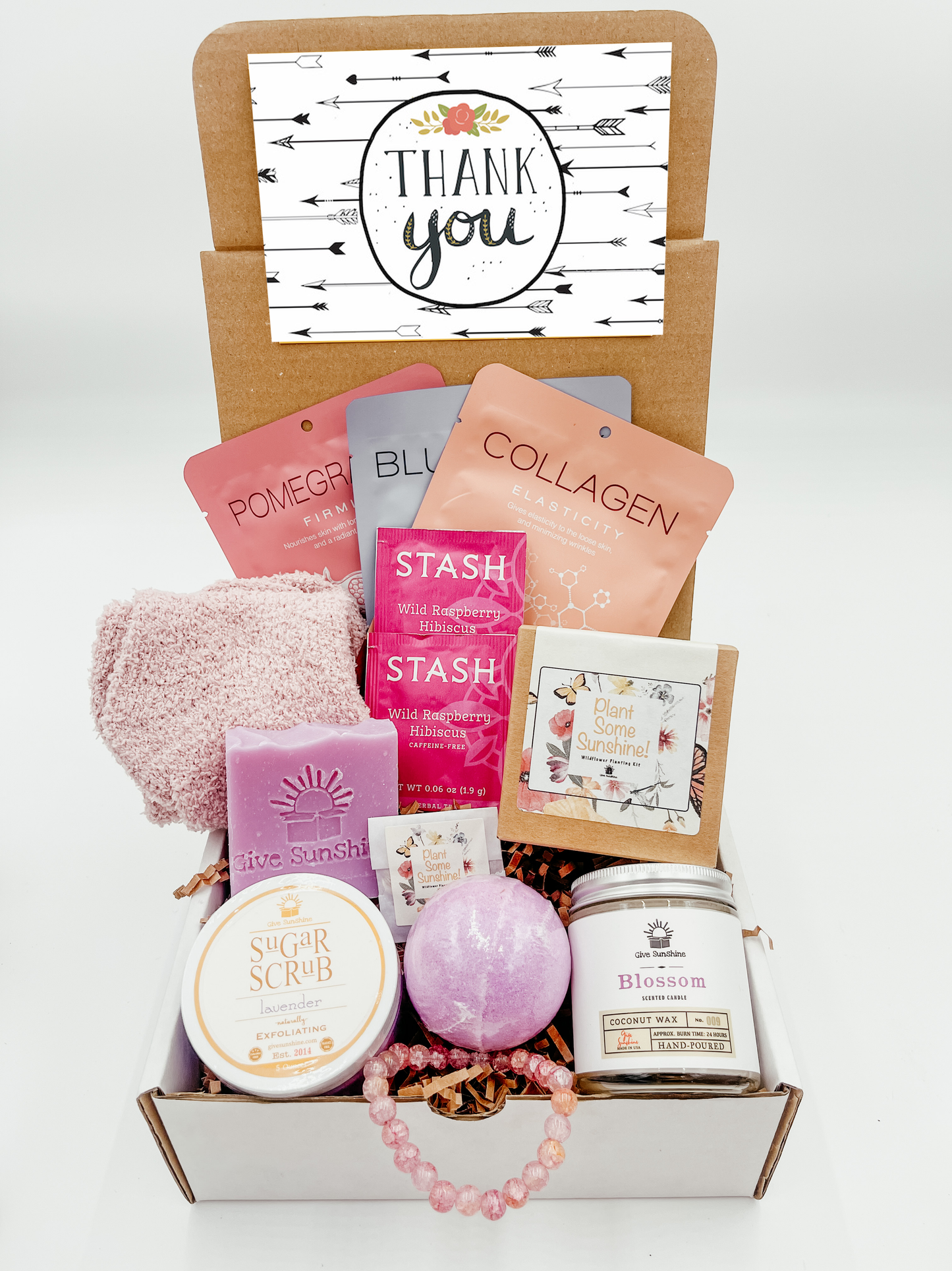 A gift box with various pink and purple colored items including a glass beaded bracelet, comfy socks, a blossoms-scented candle, lavender sugar scrub, a bath bomb, lavender goats milk soap, a wildflower kit, and a packet of wild raspberry hibiscus tea.