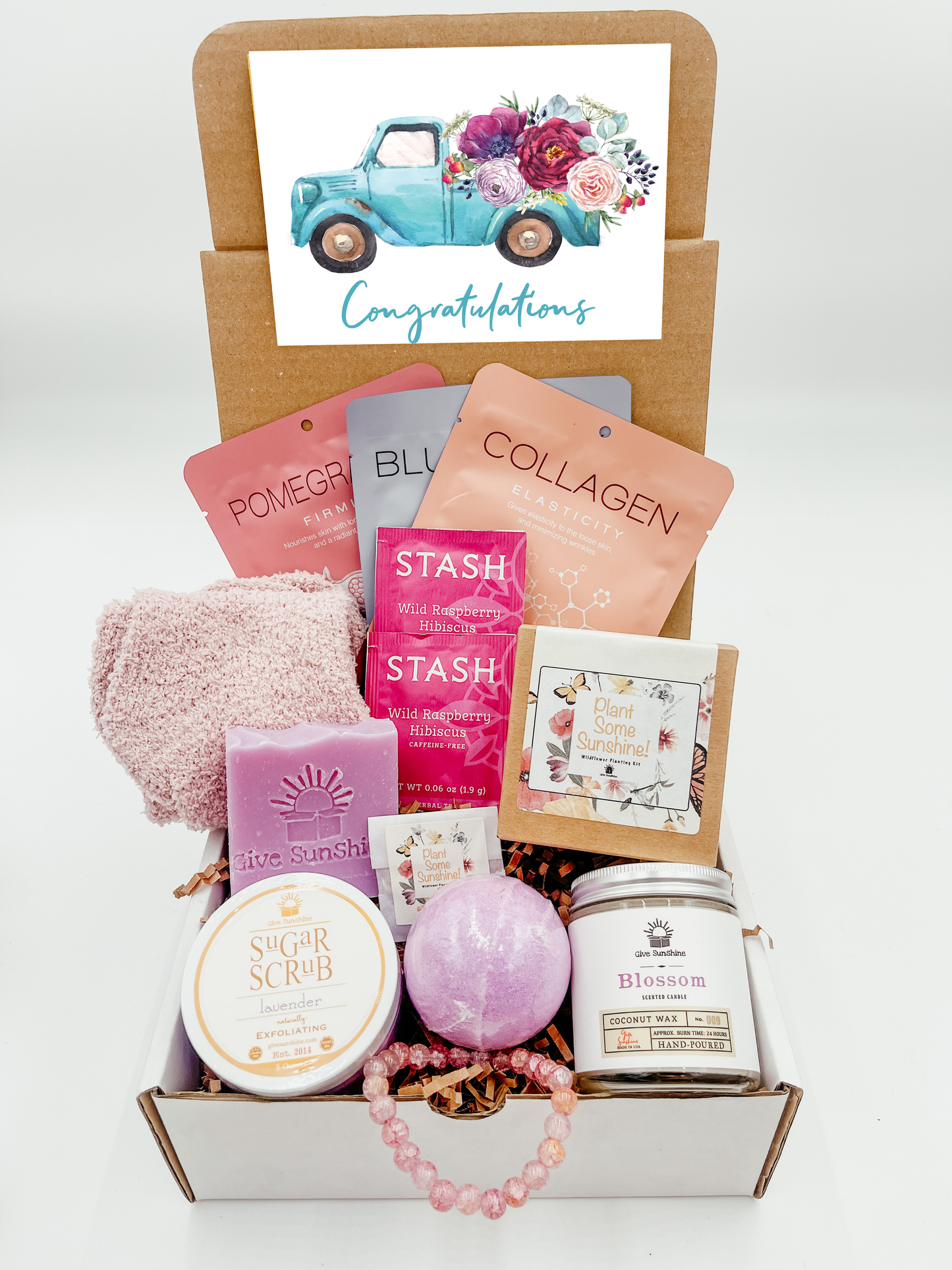 A gift box with various pink and purple colored items including a glass beaded bracelet, comfy socks, a blossoms-scented candle, lavender sugar scrub, a bath bomb, lavender goats milk soap, a wildflower kit, and a packet of wild raspberry hibiscus tea.
