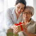 Smiling woman giving a gift box with a red ribbon to an elderly woman as they share a warm, joyful moment together.
