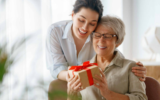 Smiling woman giving a gift box with a red ribbon to an elderly woman as they share a warm, joyful moment together.