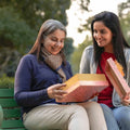 Two women sitting on a park bench smiling as one opens a decorative gift box, sharing a warm and joyful moment outdoors.