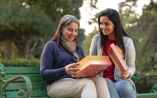 Two women sitting on a park bench smiling as one opens a decorative gift box, sharing a warm and joyful moment outdoors.