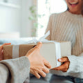Close-up of a person's hands giving a gift wrapped in brown paper and a white ribbon to a smiling recipient.