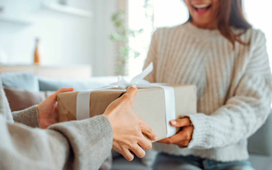 Close-up of a person's hands giving a gift wrapped in brown paper and a white ribbon to a smiling recipient.